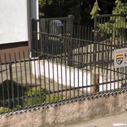 Kornel Ogrody - Metalowe ogrodzenie posesji z furtką, widok z zewnątrz. Na ogrodzeniu tabliczka informacyjna. W tle zieleń drzew i krzewów.