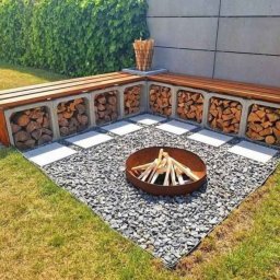 Kornel Ogrody - Ogród z miejscem na ognisko: betonowe siedzisko z drewnem, żwirowe podłoże i palenisko z drewnem. Tło stanowi zielony żywopłot i betonowy mur.
