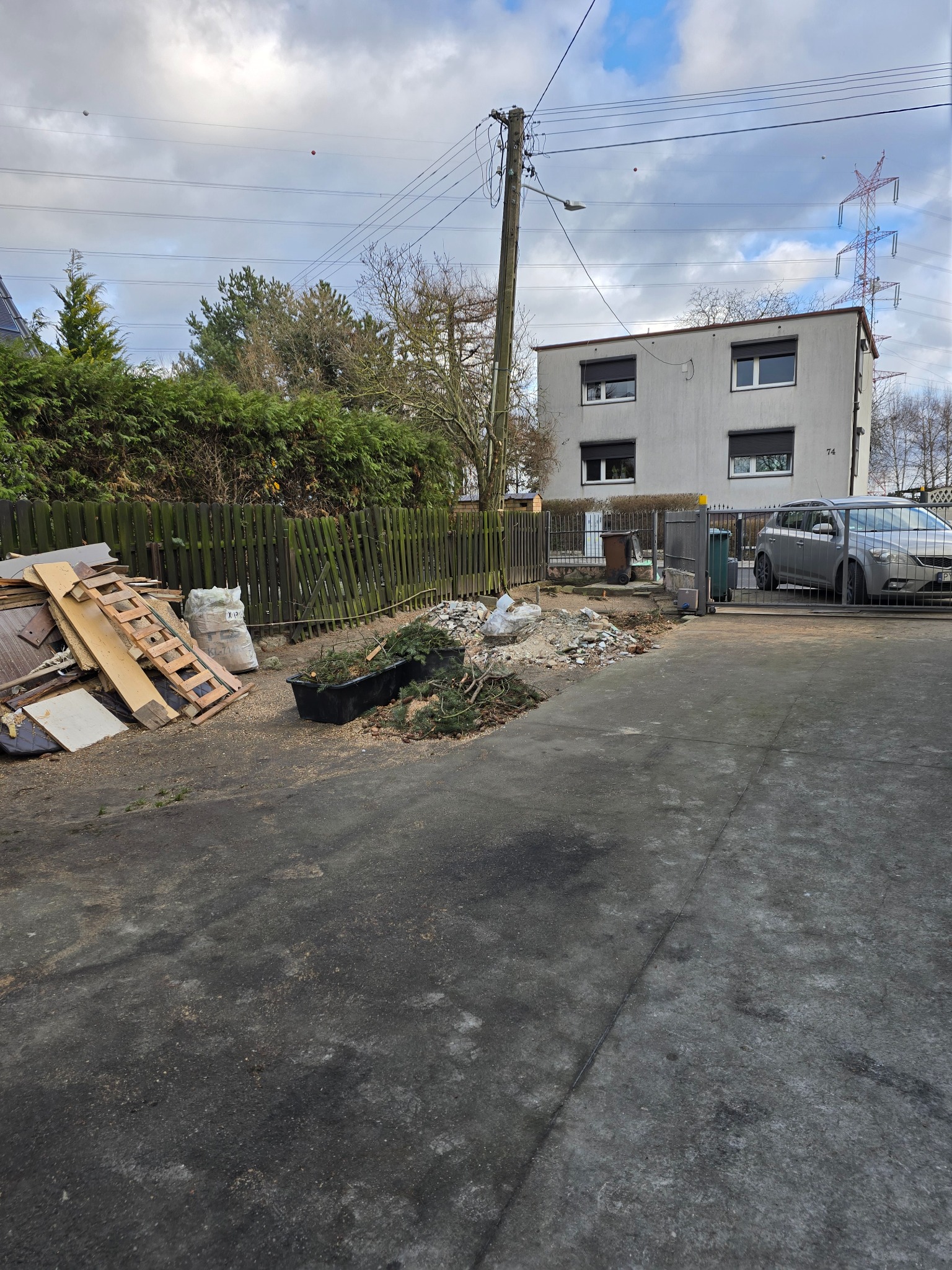 Teren po wycince drzew i krzewów: gałęzie w pojemniku, drewno, gruz, worki. Widoczny płot, budynek mieszkalny i zaparkowany samochód. Prace porządkowe w toku.
