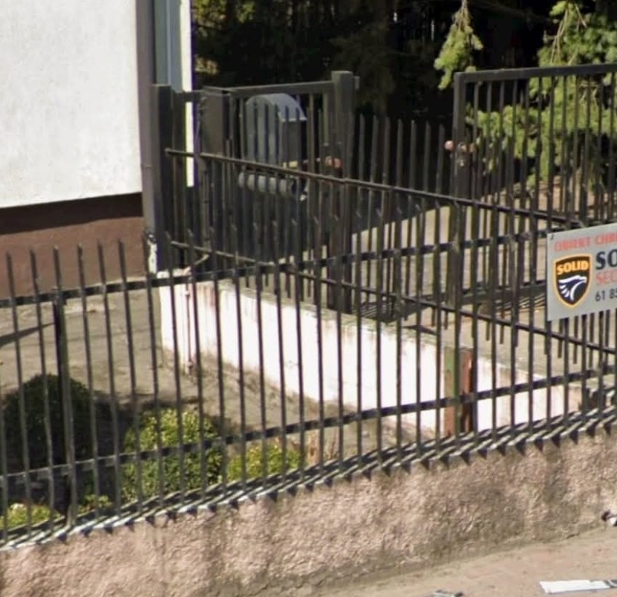 Metalowe ogrodzenie posesji z furtką, widok z zewnątrz. Na ogrodzeniu tabliczka informacyjna. W tle zieleń drzew i krzewów.