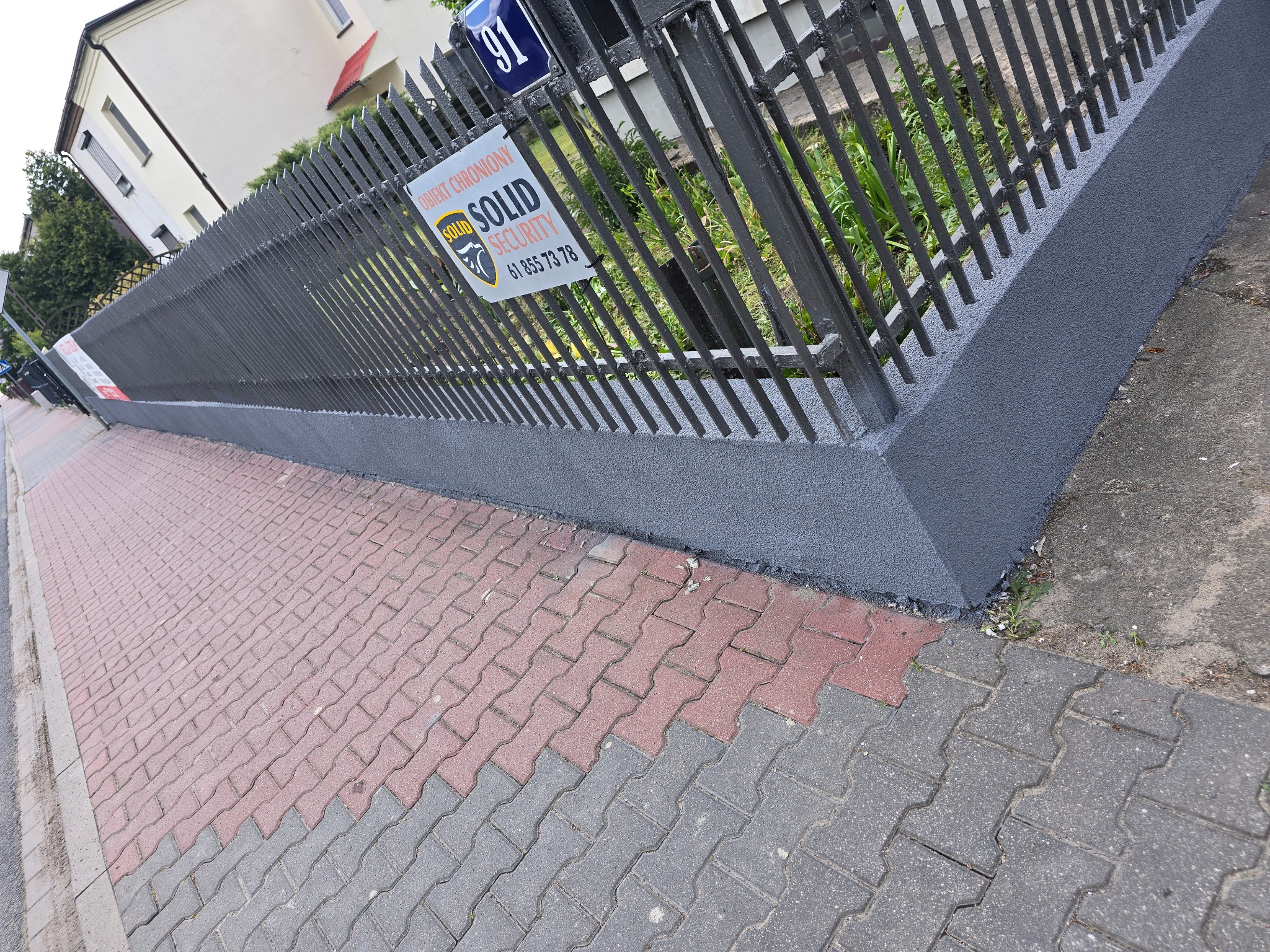 Odnowione, pomalowane na szaro metalowe ogrodzenie posesji z tabliczką 'Obiekt Chroniony SOLID Security' i numerem 91, widok z poziomu chodnika z kostki.