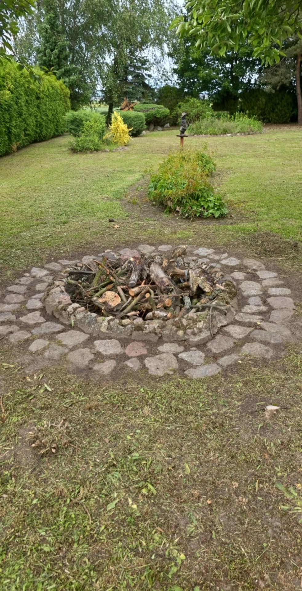 Ognisko otoczone kamieniami w ogrodzie. W tle widoczne drzewa i krzewy. Ognisko wypełnione drewnem i gałęziami, gotowe do rozpalenia. Ujęcie z perspektywy lekko z góry.