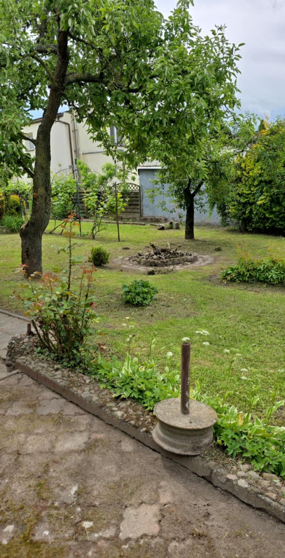 Zadbany ogród z trawnikiem, drzewami i rabatami kwiatowymi. W tle widoczny budynek. Na pierwszym planie element dekoracyjny z metalowym słupkiem.