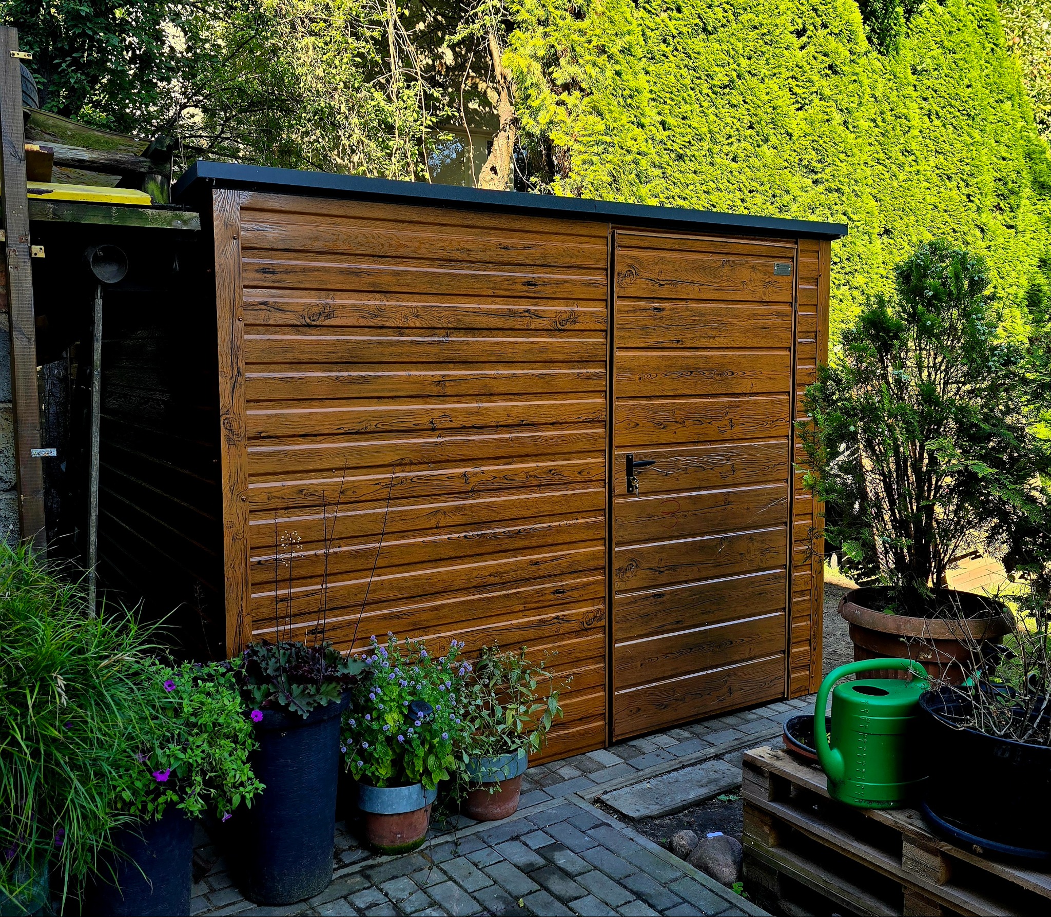 Drewniana szopa ogrodowa z czarnym dachem, otoczona zielenią i kwiatami w donicach. Widoczna konewka i kostka brukowa. Ciepłe światło słoneczne.