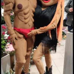 Ewelina Polus - Dwie osoby w kostiumach: mężczyzna w stroju kulturysty i kobieta w czarnej sukience z frędzlami, stoją obok siebie na tle kwiatów.