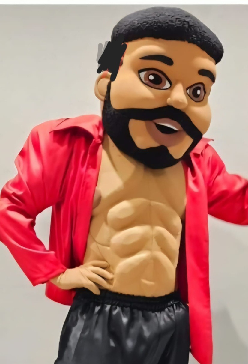 Męska maskotka z odsłoniętym torsem i czerwoną koszulą, pozująca na jasnym tle. Nietypowy kostium z widocznymi mięśniami brzucha i czarną brodą.