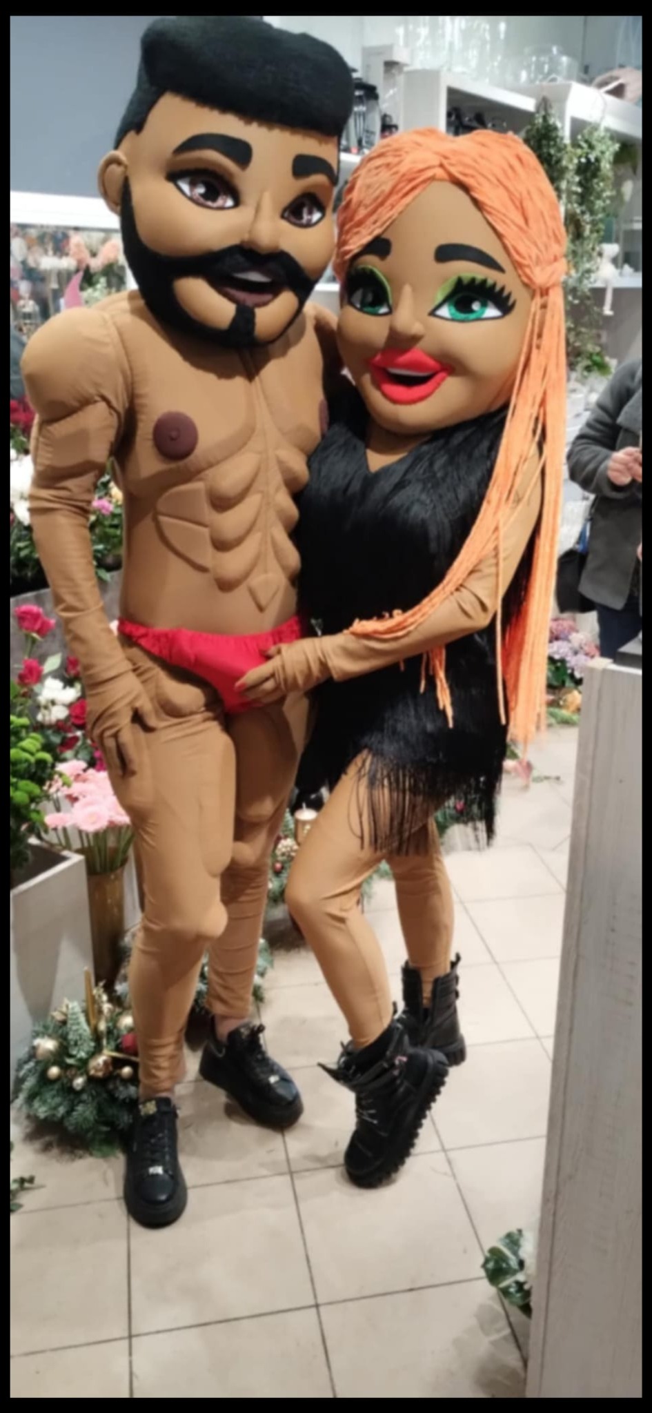 Dwie osoby w kostiumach: mężczyzna w stroju kulturysty i kobieta w czarnej sukience z frędzlami, stoją obok siebie na tle kwiatów.