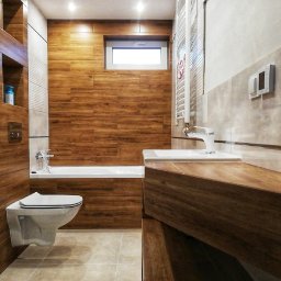 USŁUGI REMONTOWO-BUDOWLANE ROBERT JAMROZ - Nowoczesna łazienka z drewnopodobnymi płytkami na ścianach i podłodze. Umywalka z szafką z drewna, wisząca toaleta i grzejnik drabinkowy. Widoczne oświetlenie sufitowe.