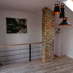USŁUGI REMONTOWO-BUDOWLANE ROBERT JAMROZ - Stylowe wnętrze z kolumną z cegły, drewnianą podłogą i nowoczesnym oświetleniem. Na ścianie obraz z motywem schodów i pianina. Widoczna barierka z drewnianym blatem.