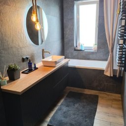 USŁUGI REMONTOWO-BUDOWLANE ROBERT JAMROZ - Nowoczesna łazienka z szarą płytką, drewnianym blatem pod umywalką i okrągłym lustrem z podświetleniem. Uwagę przykuwa lampa wisząca na sznurze.