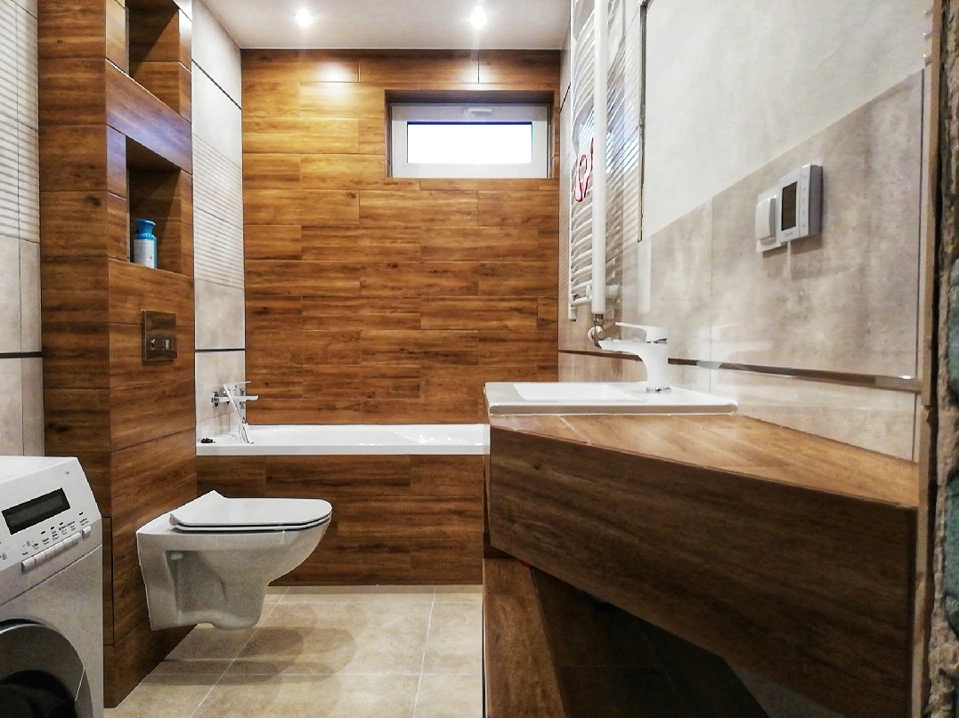 Nowoczesna łazienka z drewnopodobnymi płytkami na ścianach i podłodze. Umywalka z szafką z drewna, wisząca toaleta i grzejnik drabinkowy. Widoczne oświetlenie sufitowe.