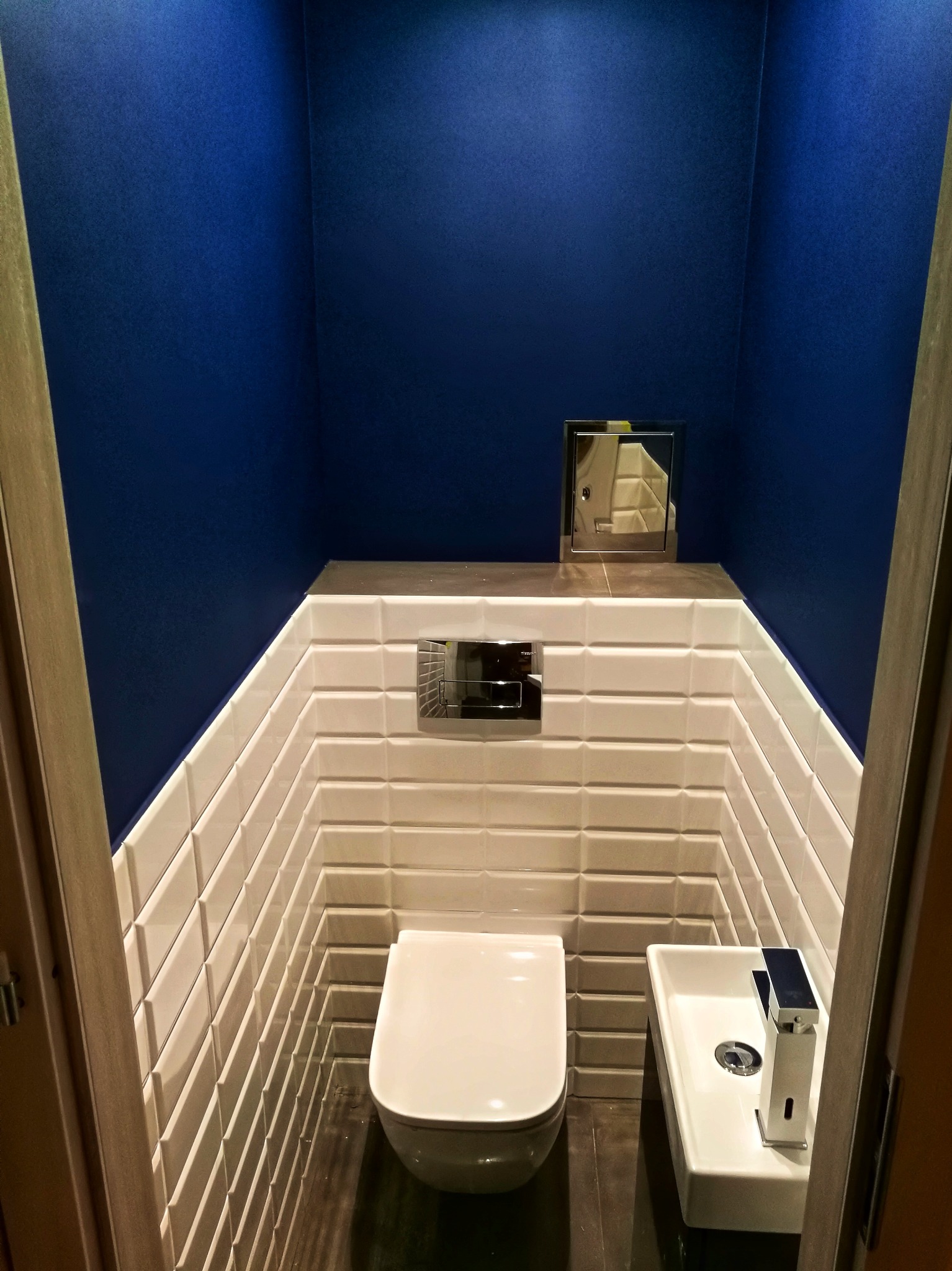 Kompaktowa toaleta w odcieniach bieli i granatu. Uwagę przykuwają białe, wypukłe płytki ułożone w oryginalny sposób oraz minimalistyczna umywalka.