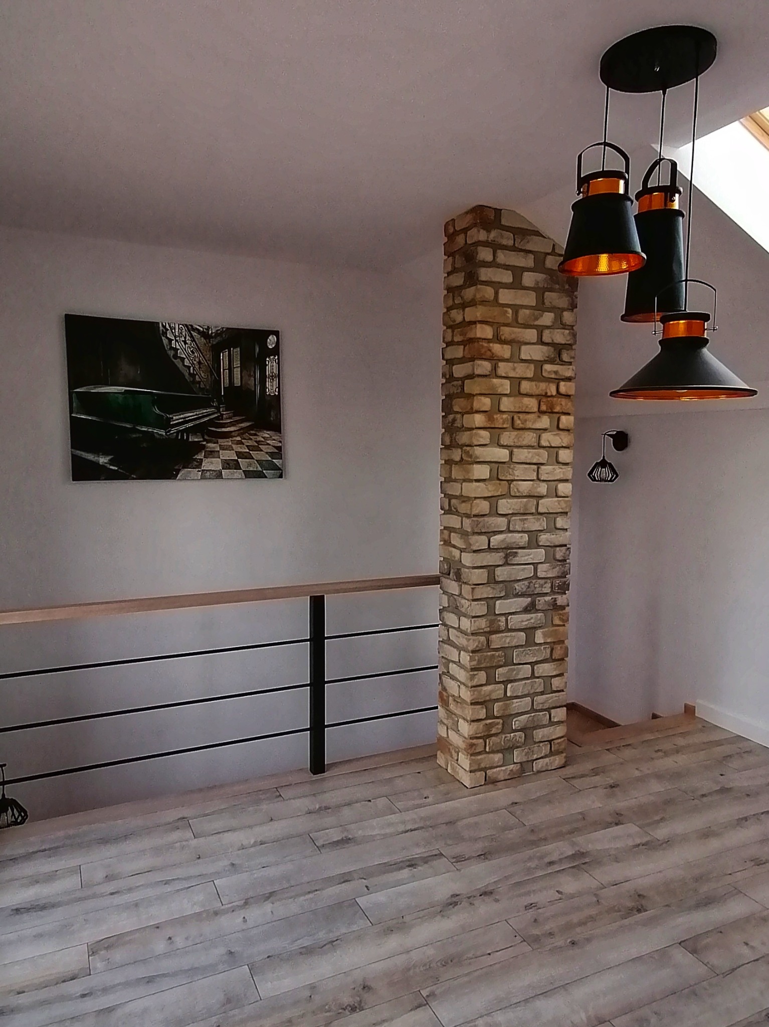 Stylowe wnętrze z kolumną z cegły, drewnianą podłogą i nowoczesnym oświetleniem. Na ścianie obraz z motywem schodów i pianina. Widoczna barierka z drewnianym blatem.