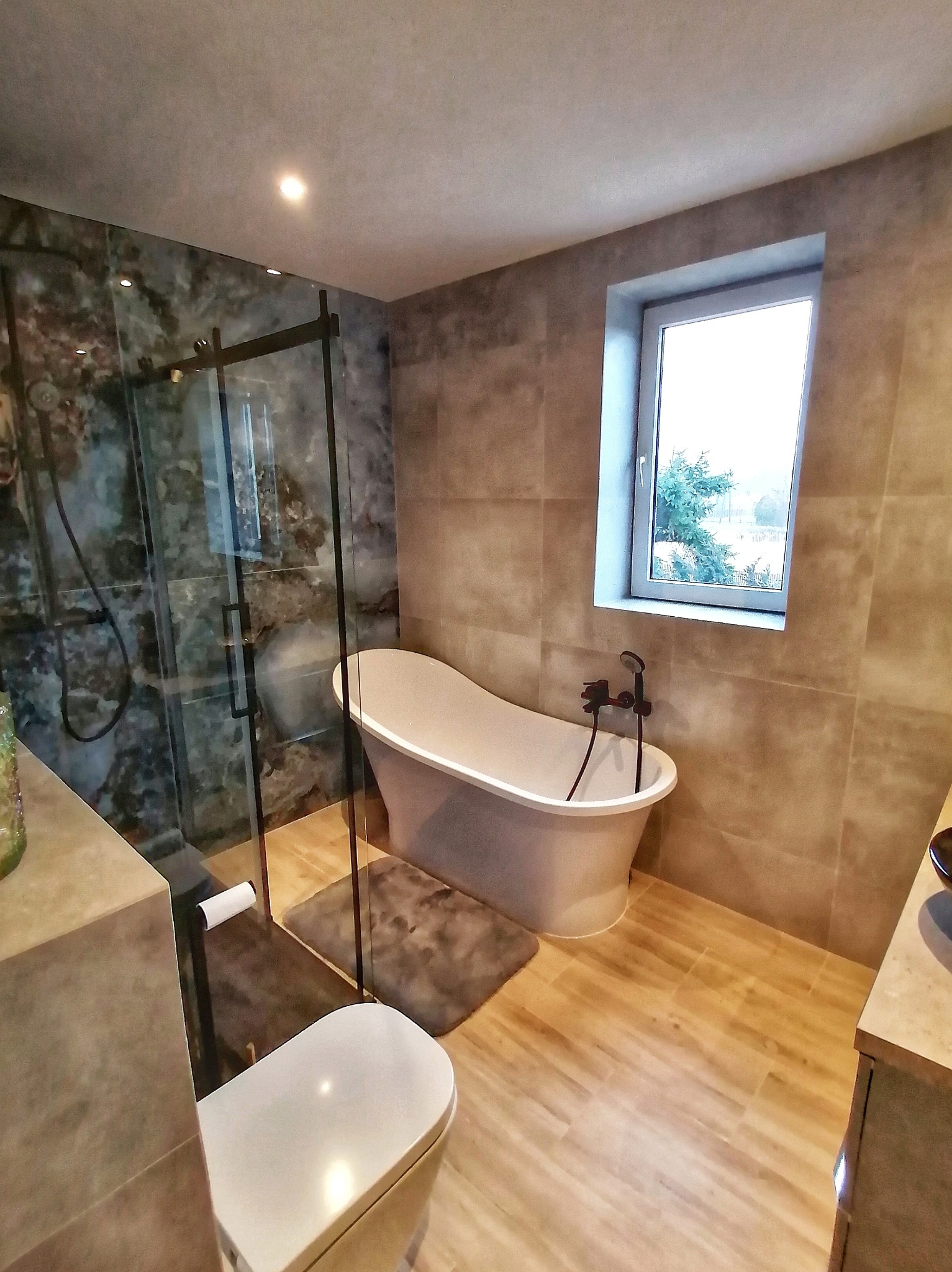 Nowoczesna łazienka z wolnostojącą wanną, kabiną prysznicową typu walk-in z czarnymi ramami i drewnopodobną podłogą. Okno doświetla wnętrze.