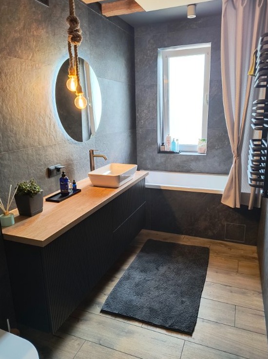 Nowoczesna łazienka z szarą płytką, drewnianym blatem pod umywalką i okrągłym lustrem z podświetleniem. Uwagę przykuwa lampa wisząca na sznurze.