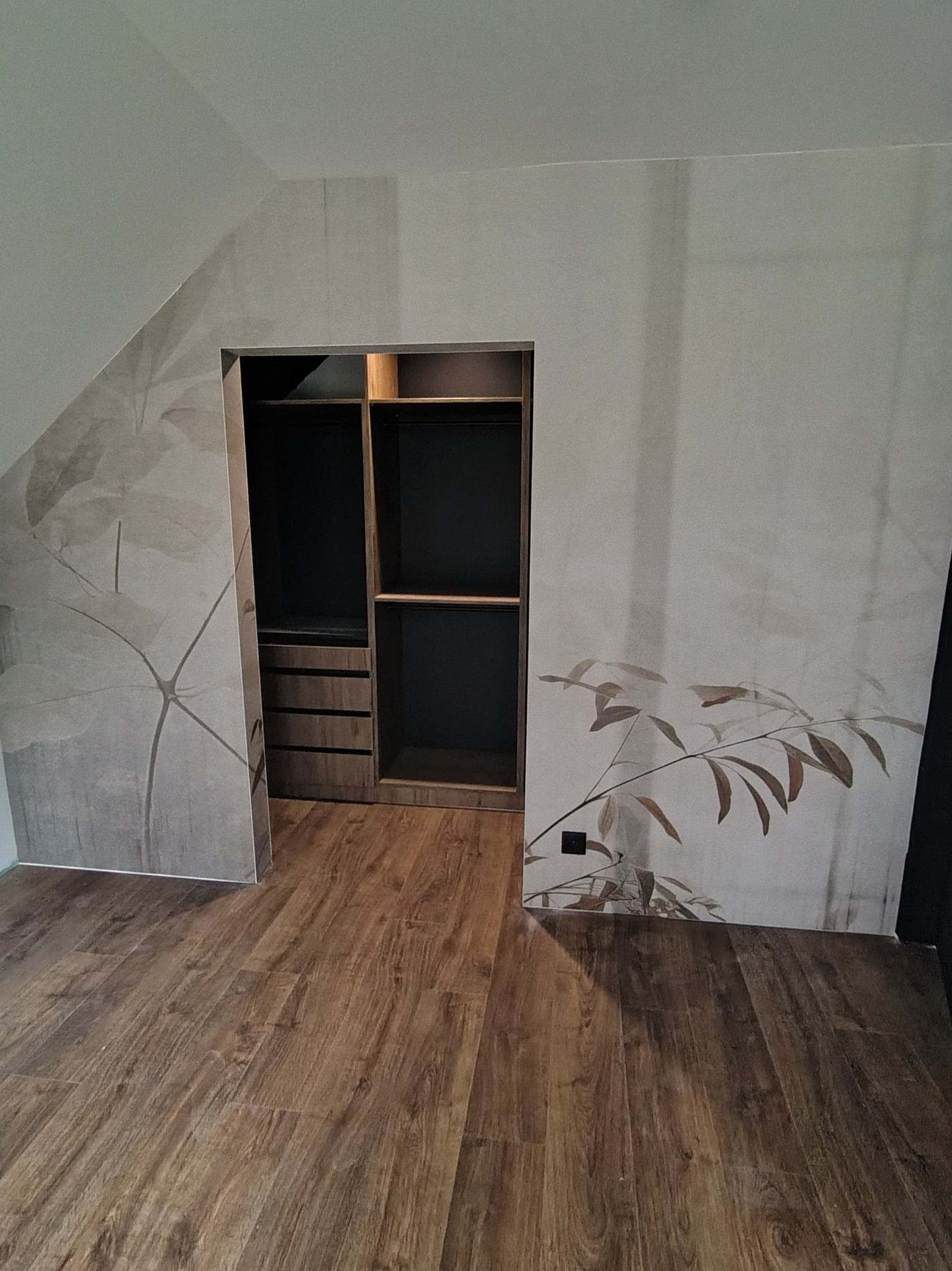 Wnęka z zabudową szafy w pokoju na poddaszu, wykończona drewnem i tapetą z motywem roślinnym. Widoczna podłoga z paneli imitujących drewno.