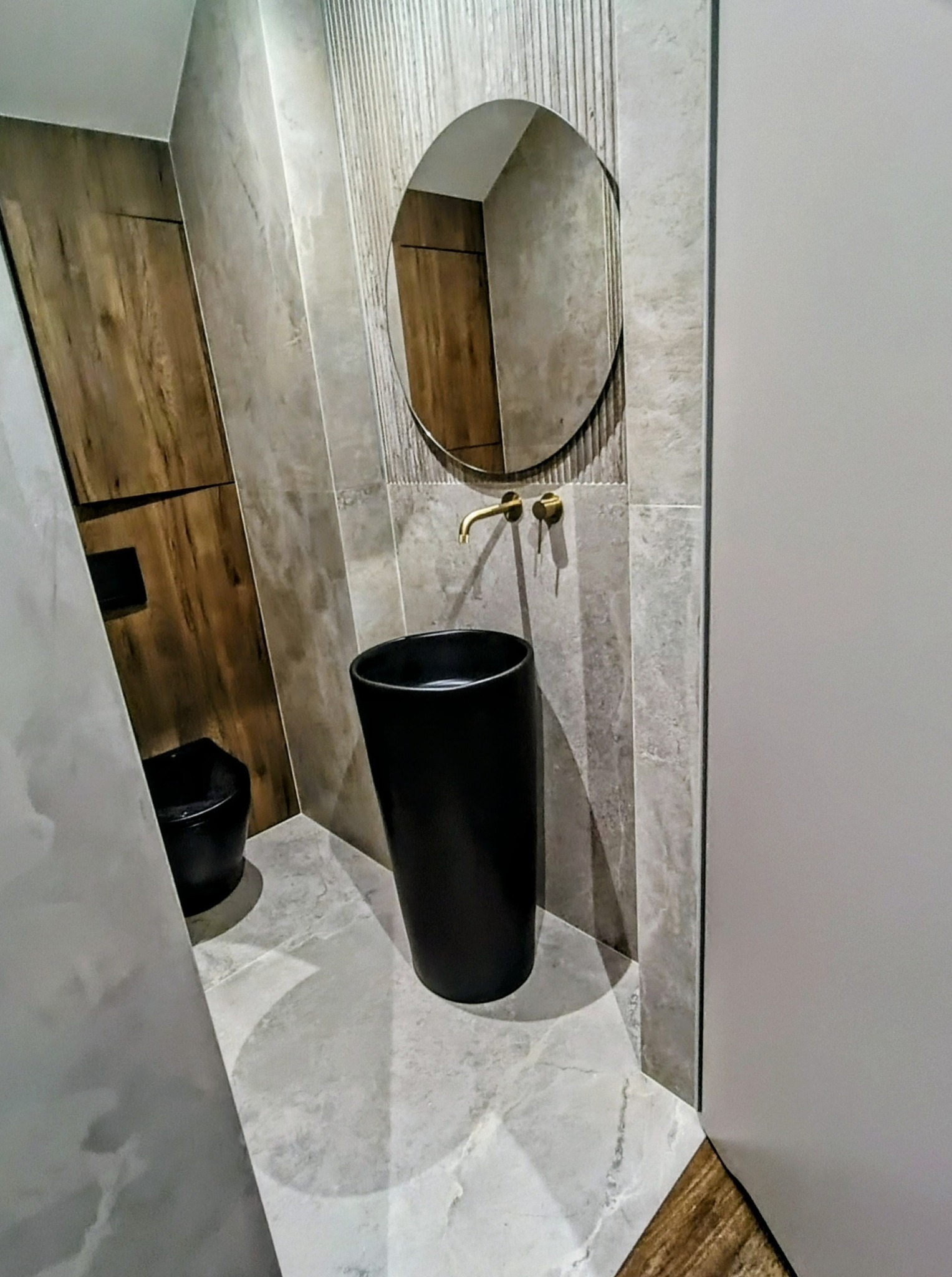 Nowoczesna łazienka z czarną umywalką wolnostojącą, okrągłym lustrem i płytkami imitującymi marmur. Widoczna toaleta w tle i złota armatura.