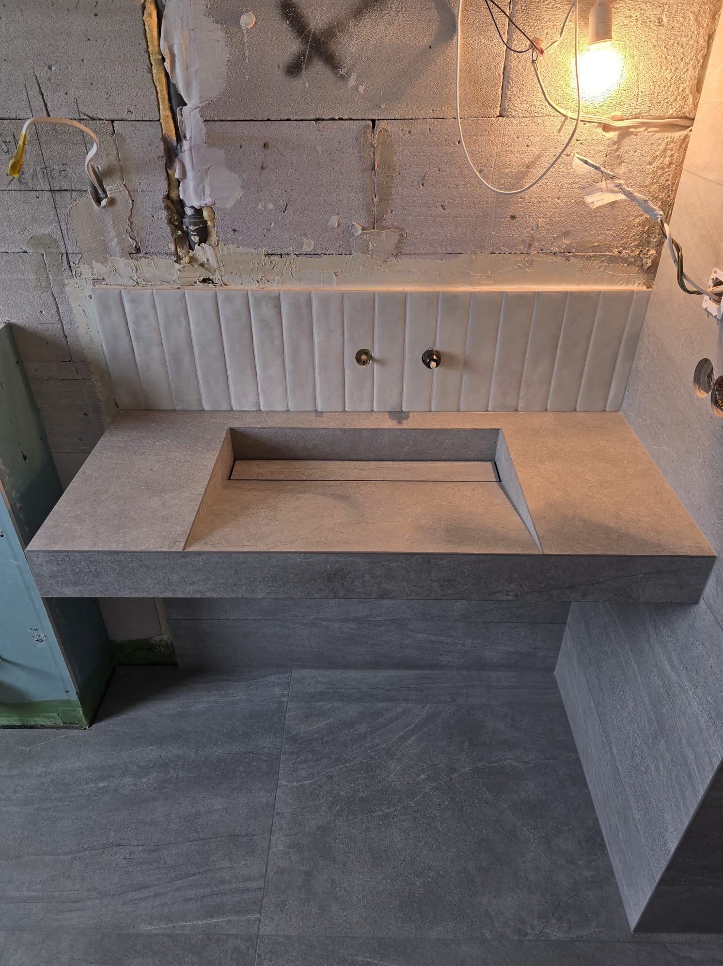 Nowoczesna łazienka w trakcie wykończenia: szara, kamienna umywalka z odpływem liniowym, płytki ścienne z subtelnym wzorem, surowy beton w tle, widoczne instalacje.