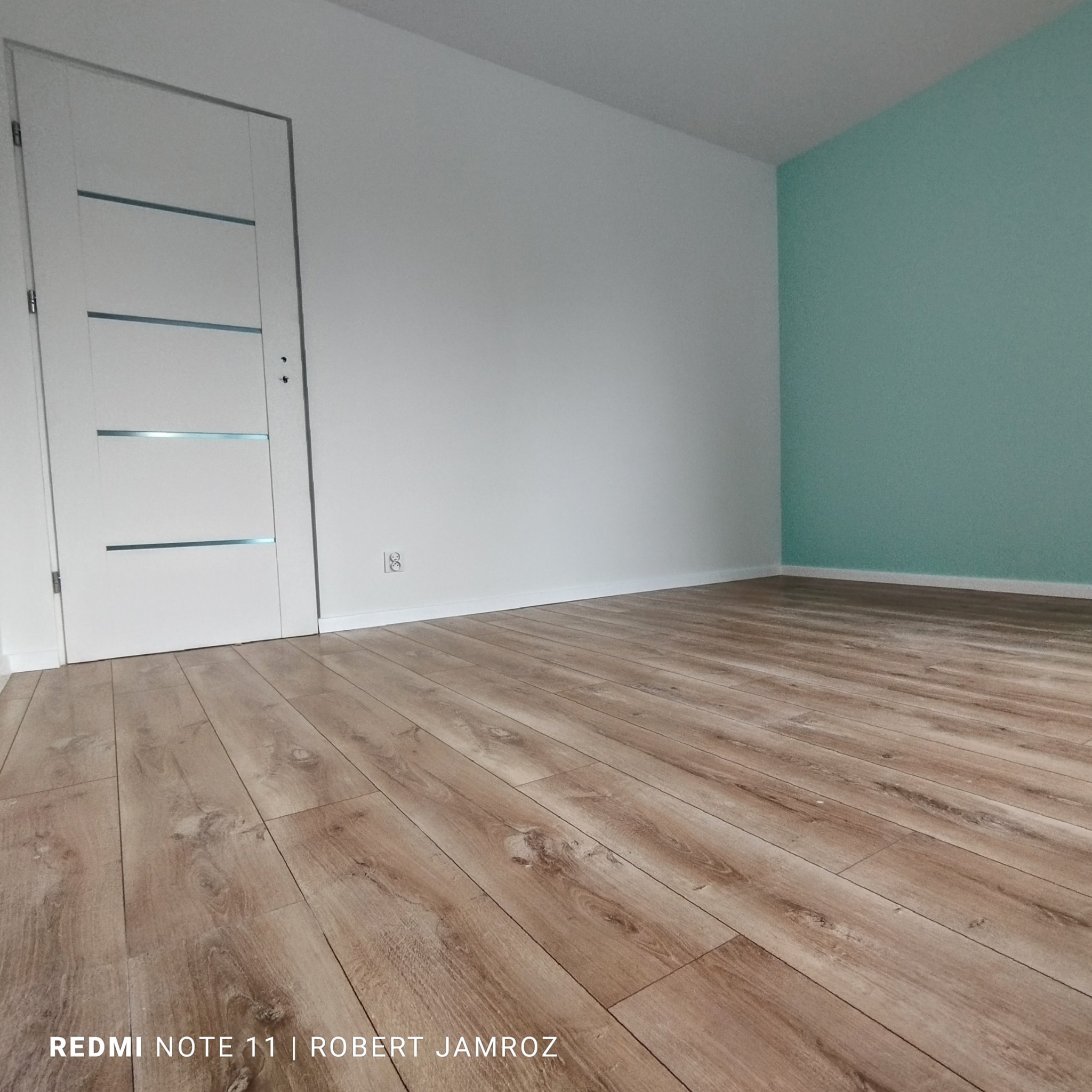 Jasne panele podłogowe w nowym pokoju z białą ścianą i turkusowym akcentem. Białe drzwi z przeszkleniami po lewej stronie. Widok z niskiej perspektywy.