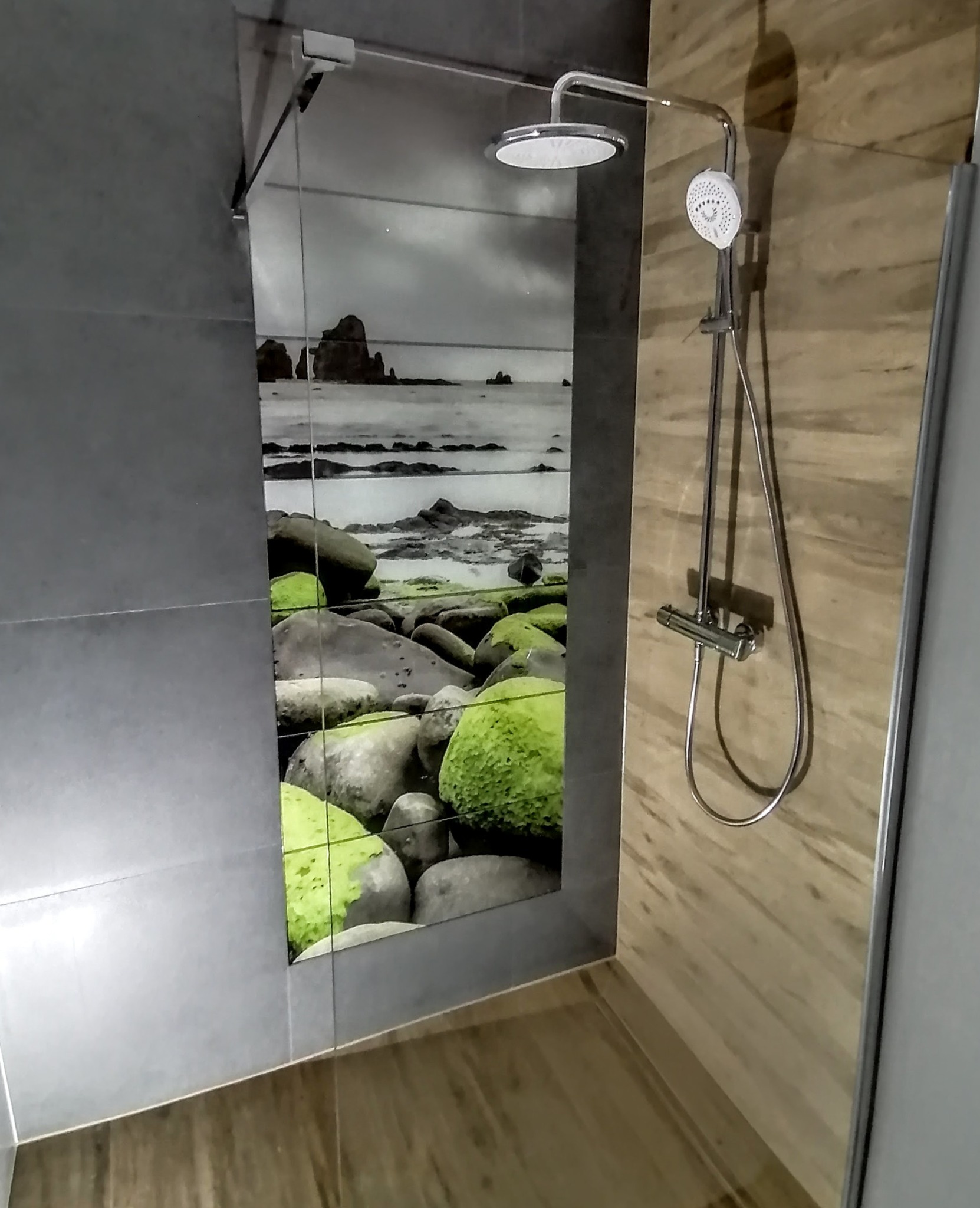 Nowoczesna kabina prysznicowa z szarymi płytkami i panelem dekoracyjnym z motywem morskiego krajobrazu. Widoczna deszczownica i bateria. Wykończenie łazienki w jasnych odcieniach.