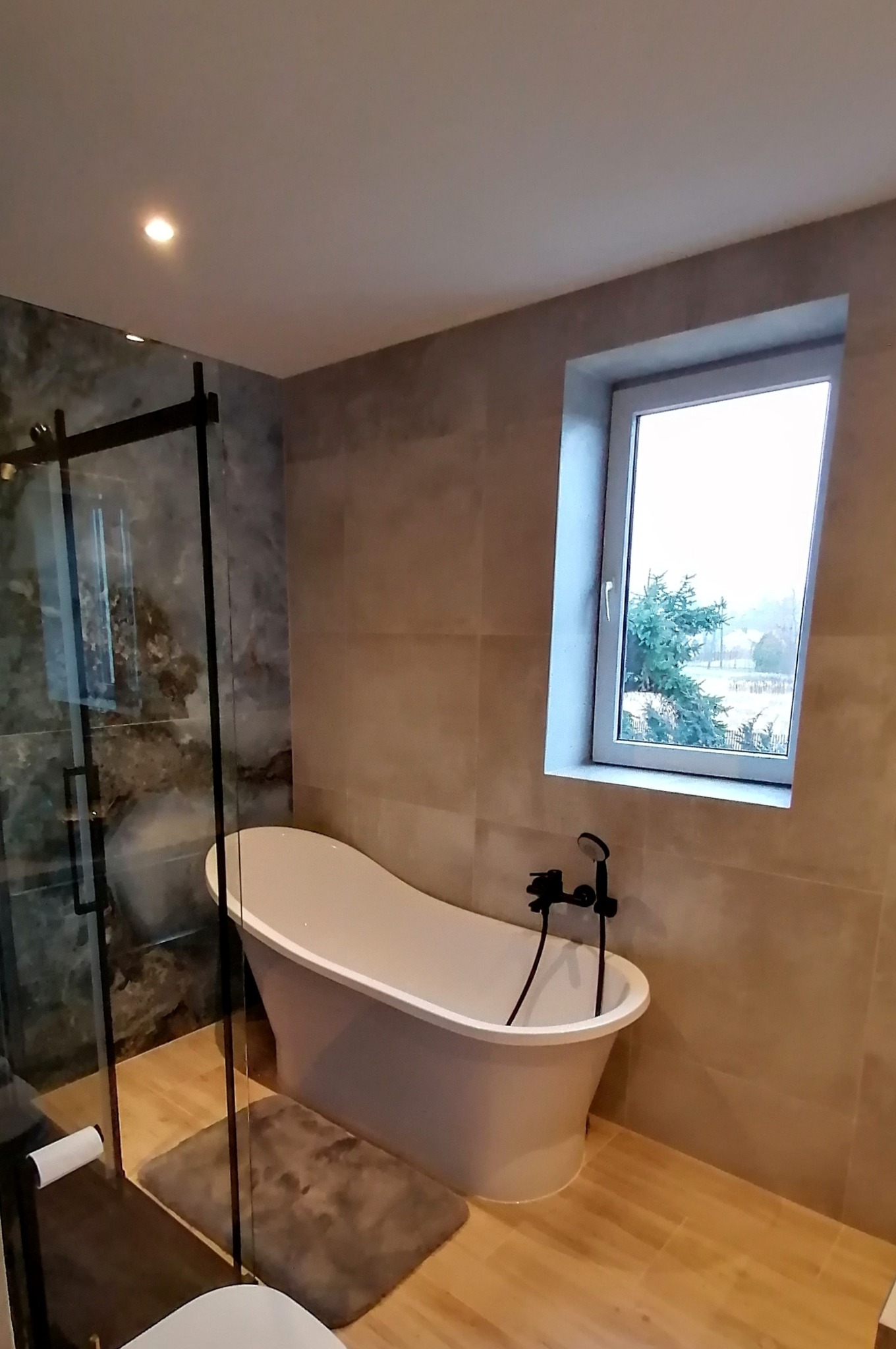 Nowoczesna łazienka w Soninie z wolnostojącą wanną, kabiną prysznicową typu walk-in i oknem z widokiem na zewnątrz. Beżowe płytki i drewnopodobna podłoga.