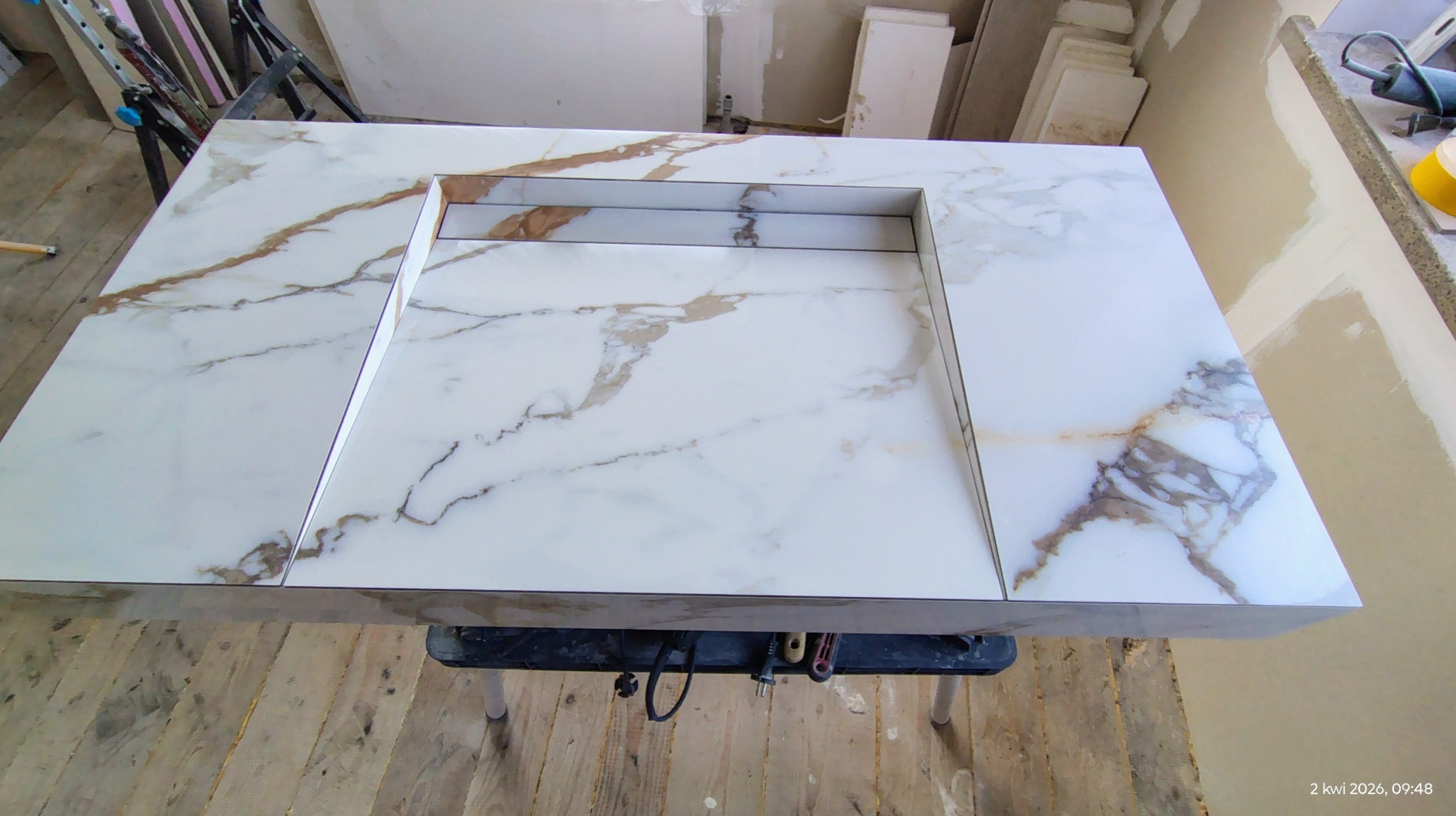 Biały blat z marmurowym wzorem, z wyciętym otworem na zlew, ustawiony na stojaku. W tle widoczne ściany w trakcie prac wykończeniowych.