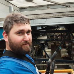 Bohdan Onyshchenko - Elektryk z brodą trzyma rury w hali produkcyjnej z metalową konstrukcją i przezroczystym dachem. Widok z góry na magazyn z materiałami.