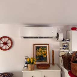 Air-Tech Piotr Dębek - Biały klimatyzator na ścianie w pokoju z dekoracjami w stylu vintage: obraz, kwiaty, zegar. Wyświetlacz klimatyzatora pokazuje 30 stopni.