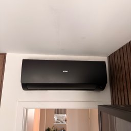Air-Tech Piotr Dębek - Czarna klimatyzacja Haier zamontowana na białej ścianie nad drzwiami. Widok na wnętrze pokoju z rośliną i meblami. Minimalistyczny design.