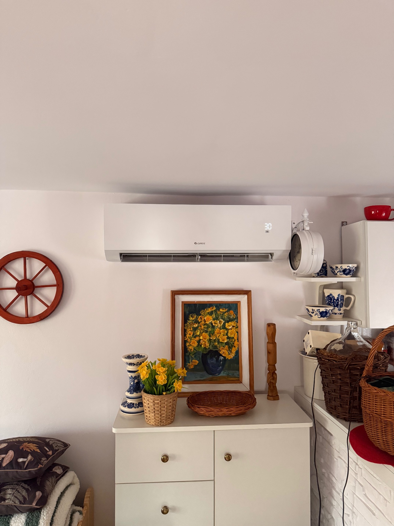 Biały klimatyzator na ścianie w pokoju z dekoracjami w stylu vintage: obraz, kwiaty, zegar. Wyświetlacz klimatyzatora pokazuje 30 stopni.
