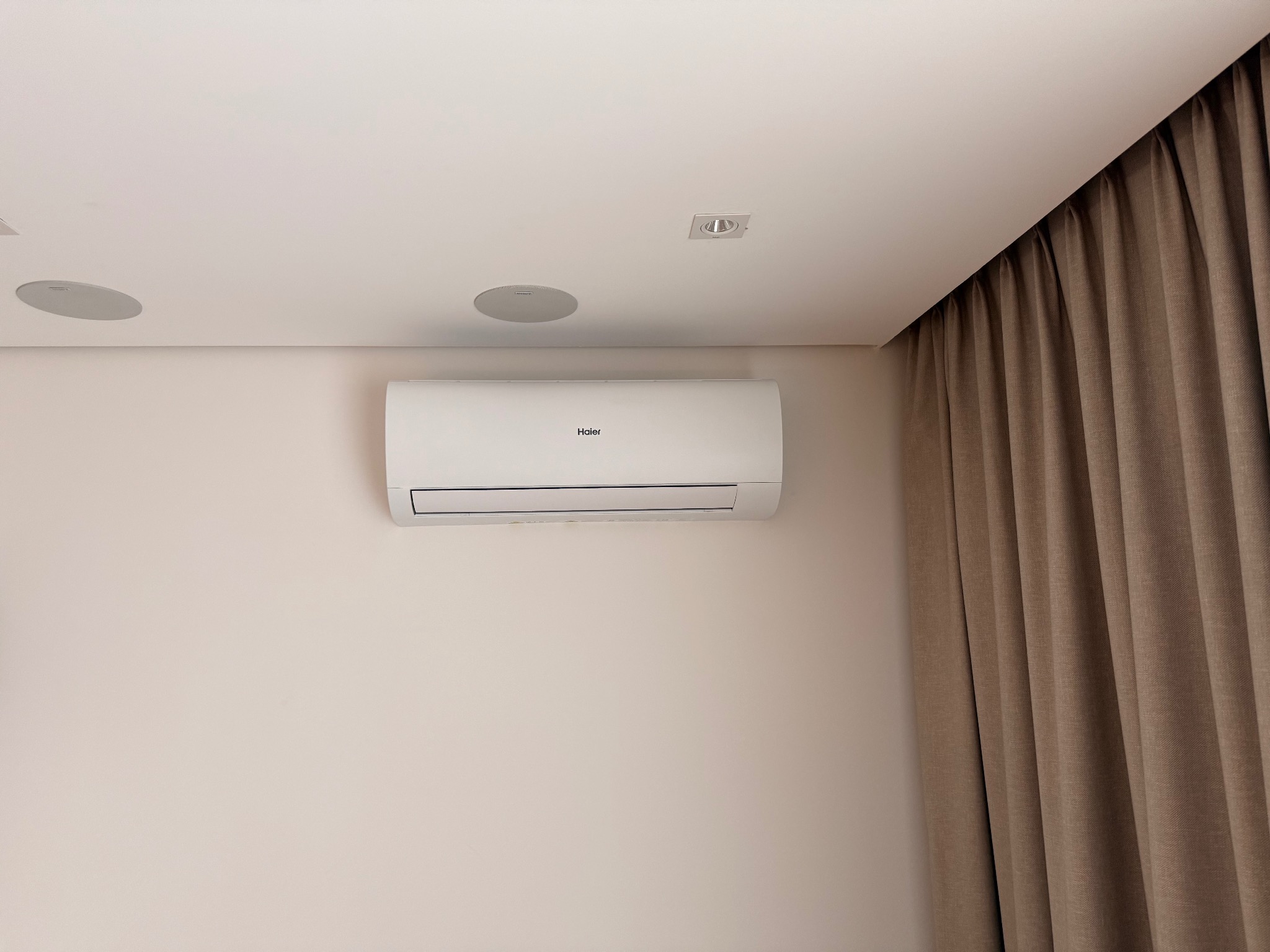 Biały klimatyzator Haier na ścianie w jasnym wnętrzu, z widocznymi głośnikami sufitowymi i zasłonami w tle. Minimalistyczny design.
