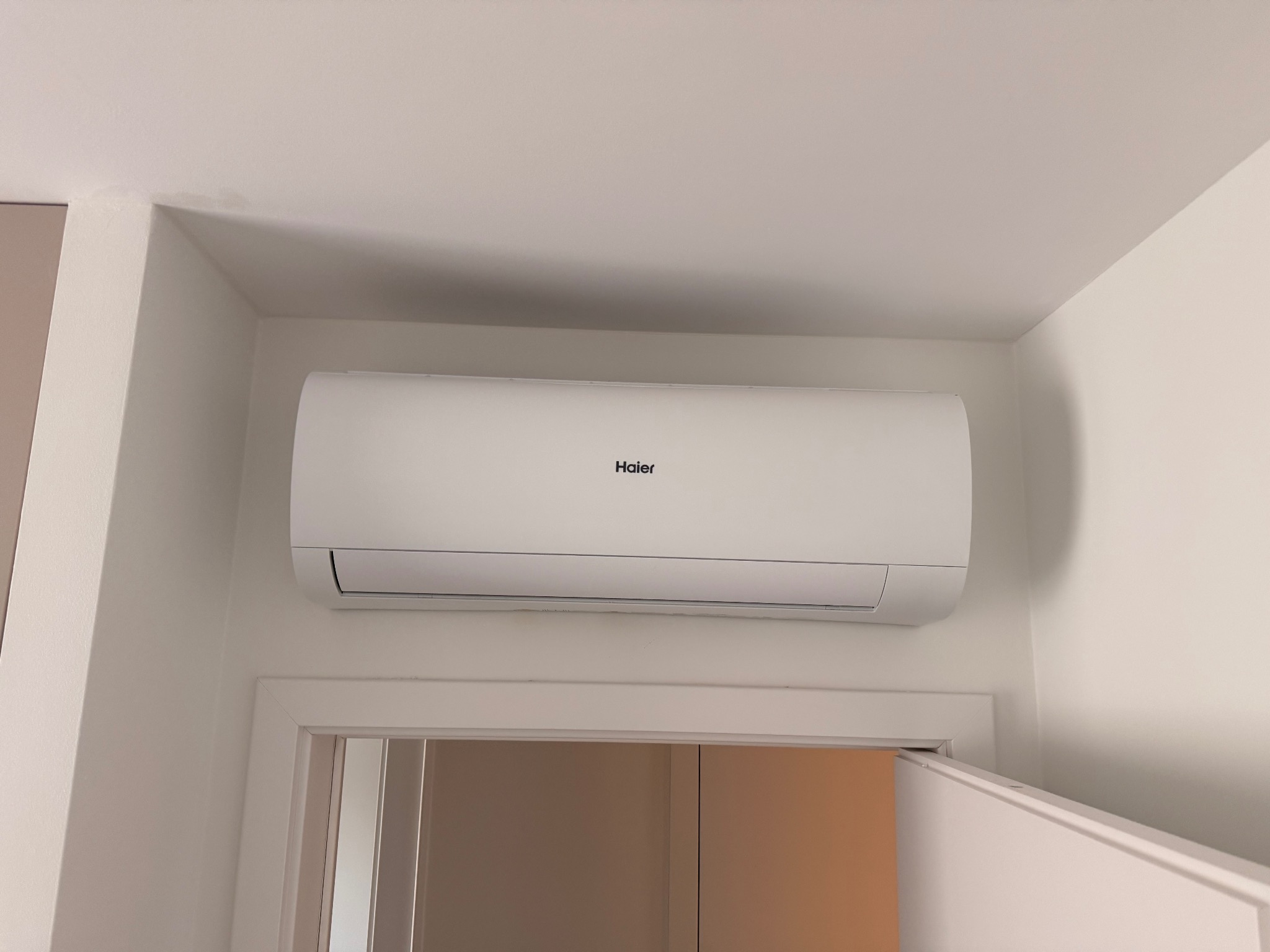 Biała klimatyzacja Haier zamontowana nad drzwiami w jasnym pomieszczeniu. Widoczny fragment ściany i sufitu. Minimalistyczny design urządzenia.