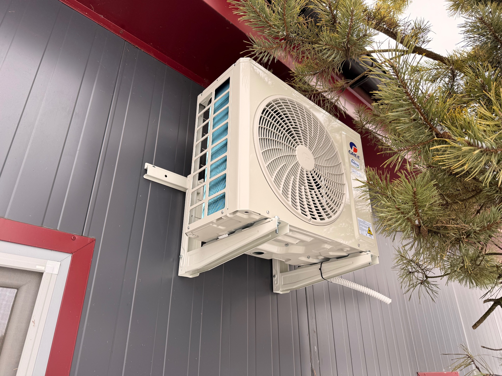 Zamontowana klimatyzacja Gree na szarej elewacji z blachy trapezowej, widoczna z boku, z fragmentem sosny w tle. Biała obudowa, niebieskie filtry.