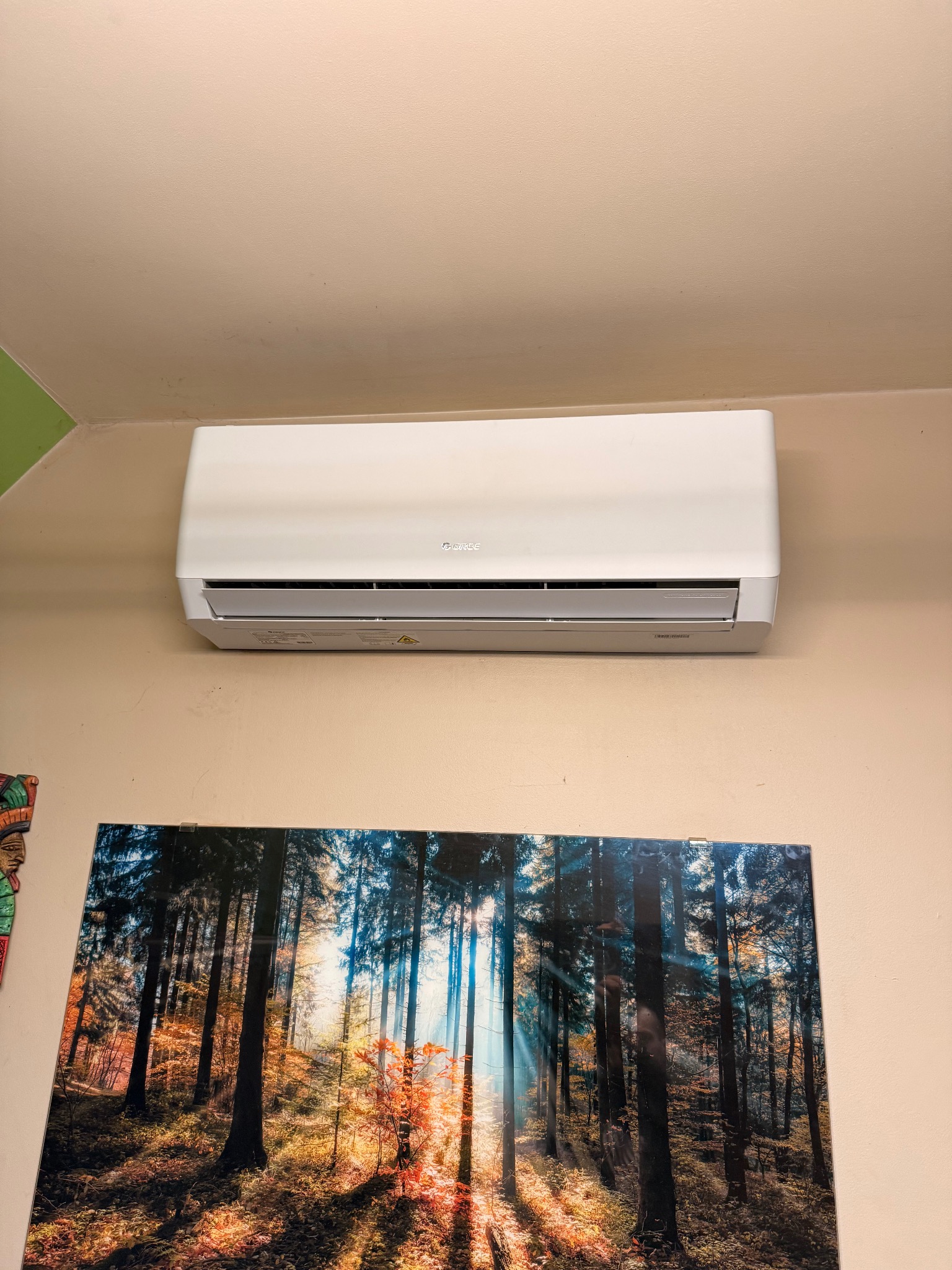Biały klimatyzator marki Gree zamontowany na ścianie nad obrazem leśnym. Widoczne detale urządzenia i otoczenia, w tym fragment dekoracji ściennej po lewej stronie.