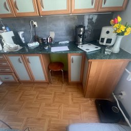 Pol - Montaż i wykończenia - Kuchnia z drewnianymi szafkami, blatem z zielonego marmuru i kwiatami w wazonie. Widoczny stół i krzesło. Na blacie ekspres do kawy, waga kuchenna i inne akcesoria.