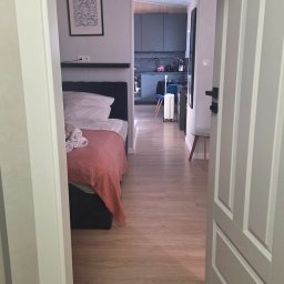 Pol - Montaż i wykończenia - Jasne wnętrze apartamentu z widokiem na sypialnię z łóżkiem i kuchnię w tle, widziane przez otwarte drzwi. Podłoga z paneli, minimalistyczny wystrój.