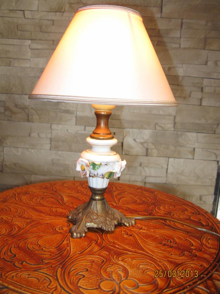 Porcelanowa lampka nocna z materiałowym kloszem, zdobiona kwiatami, stoi na okrągłym stole z wytłaczanym wzorem, prezentowana w Żmigrodzie.