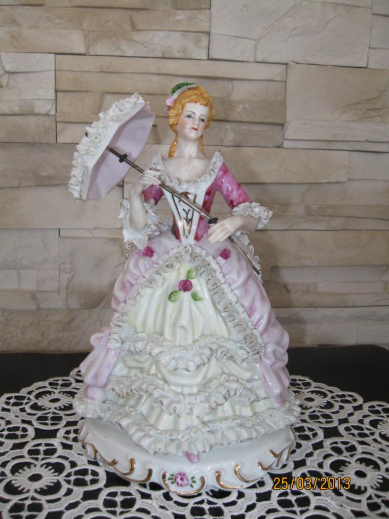 Porcelanowa figurka damy w rokokowej sukni z parasolką, stojąca na koronkowej serwetce, na tle ściany z kamienia dekoracyjnego.