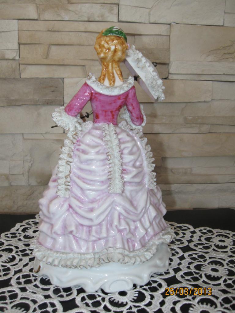 Porcelanowa figurka damy w różowej sukni z falbanami, trzymającej parasolkę, ustawiona tyłem na koronkowej serwetce, na tle ściany z kamienia dekoracyjnego.