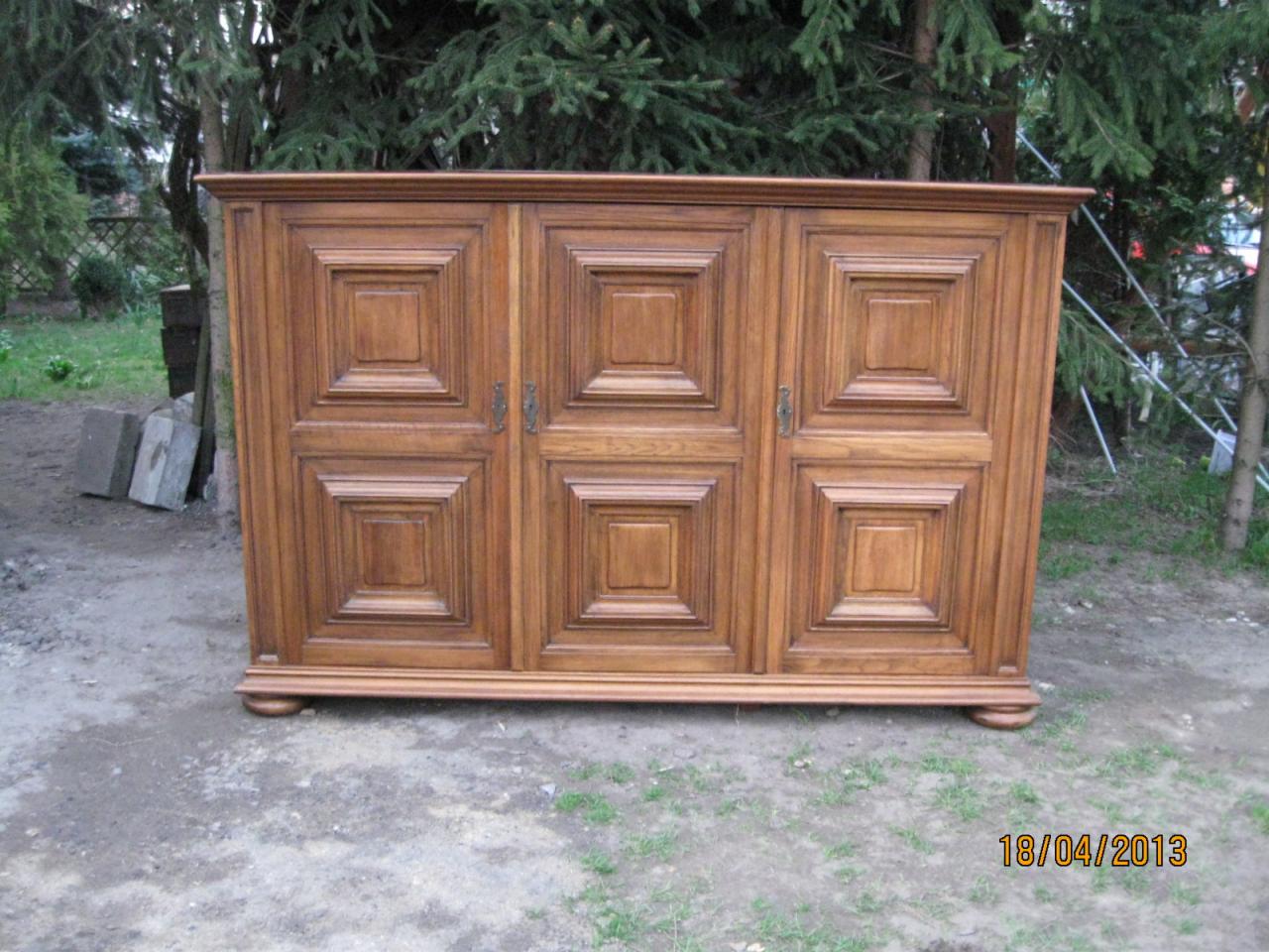 Dębowa komoda z trzema segmentami, każdy z kwadratowymi panelami, ustawiona na zewnątrz na ziemi przed zielonymi drzewami.