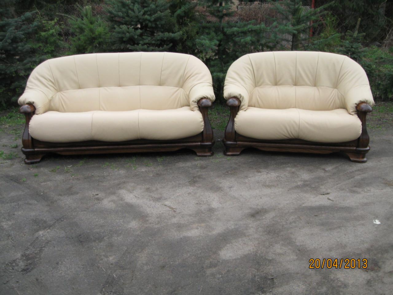 Dwa kremowe, dwuosobowe fotele tapicerowane z dębowymi stelażami, ustawione na betonowym podłożu przed zielonymi krzewami.