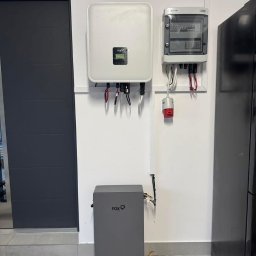 BestKlima - Zamontowany biały inwerter fotowoltaiczny Fox obok szarego akumulatora energii, z rozdzielnicą elektryczną powyżej, na tle białej ściany wewnątrz pomieszczenia.