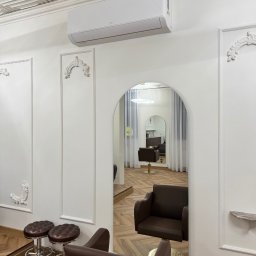 BestKlima Paulina Szczęsna - Białe wnętrze salonu z klimatyzatorem TCL na ścianie, widoczne krzesła fryzjerskie i lustro w kształcie łuku. Dekoracyjne sztukaterie na ścianach i kasetonowy sufit.