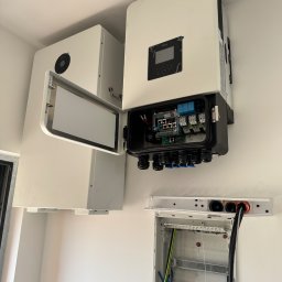 BestKlima Paulina Szczęsna - Biały inwerter fotowoltaiczny Deye z otwartą obudową, widoczne okablowanie i podłączenia, obok akumulator energii i skrzynka elektryczna na białej ścianie.