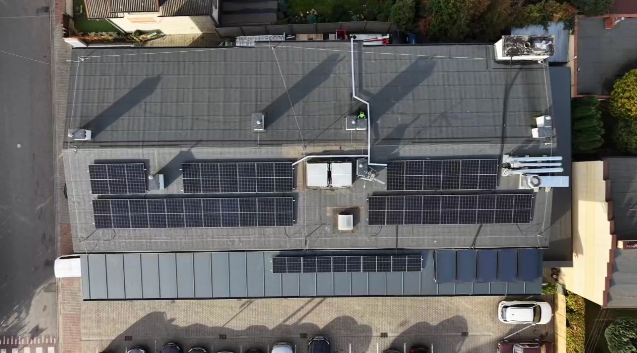 Widok z drona na dach budynku z panelami słonecznymi i klimatyzacją w Węgrowie. Instalacja fotowoltaiczna na płaskim dachu. Widoczne elementy wentylacji i zaparkowane samochody.