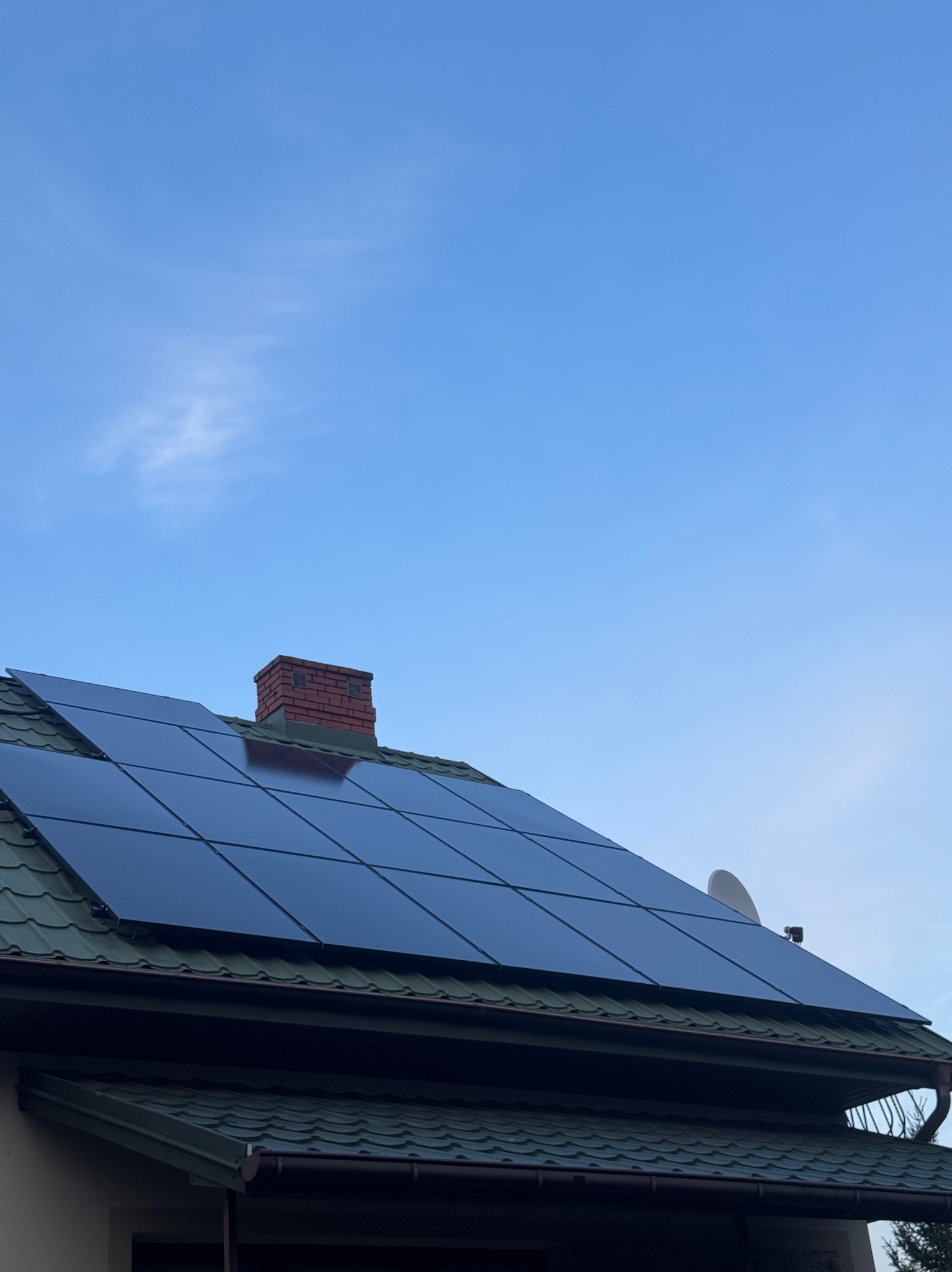 Panele fotowoltaiczne na zielonym dachu domu jednorodzinnego, widok z dołu, komin z czerwonej cegły i antena satelitarna w tle, błękitne niebo.