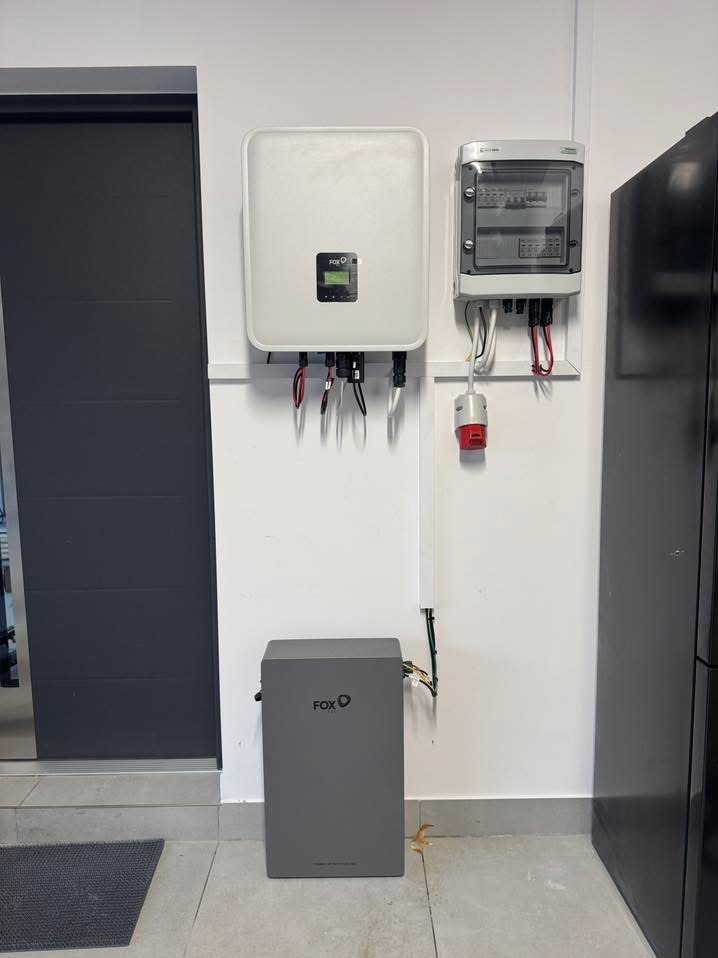 Zamontowany biały inwerter fotowoltaiczny Fox obok szarego akumulatora energii, z rozdzielnicą elektryczną powyżej, na tle białej ściany wewnątrz pomieszczenia.