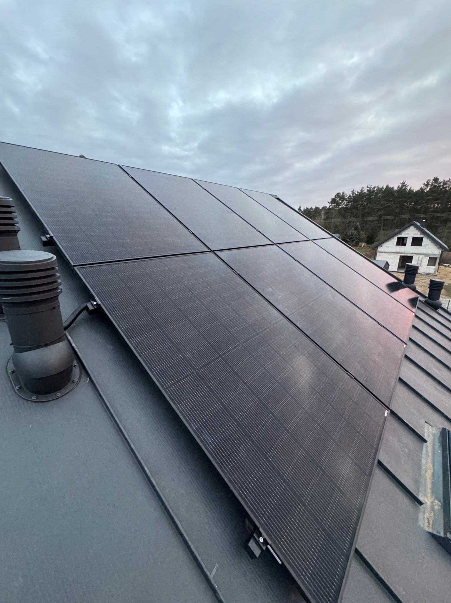 Czarny panel fotowoltaiczny na szarym dachu domu jednorodzinnego, widok z dołu pod kątem, pochmurne niebo w tle. Widoczne kominy wentylacyjne.
