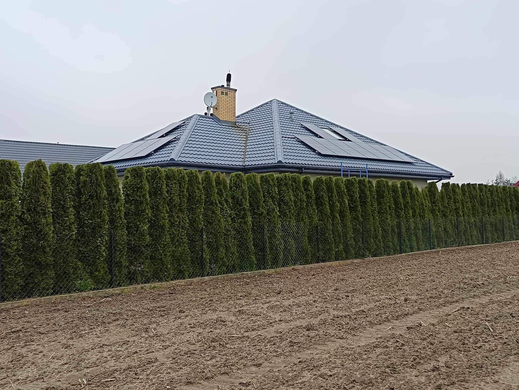 Dom jednorodzinny z szarą dachówką i panelami słonecznymi, otoczony żywopłotem i polem. Widoczny komin z anteną satelitarną. Pochmurne niebo w tle.