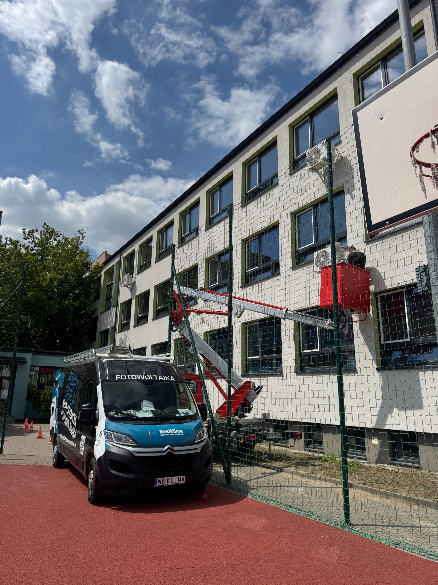 Montaż klimatyzacji na budynku szkolnym z użyciem podnośnika koszowego. Na pierwszym planie firmowy bus z logo fotowoltaiki i klimatyzacji.